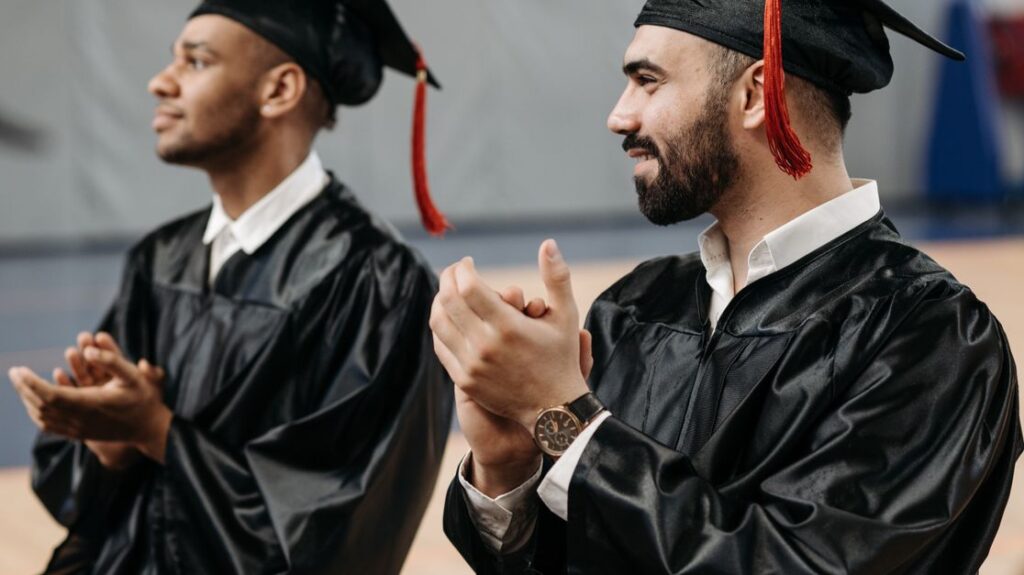 Two graduates clapping and celebrating their achievements in caps and gowns during a graduation ceremony.